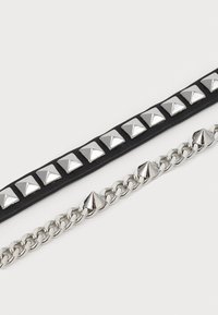 Deux accessoires : une sangle en cuir noir avec des clous en pyramide argentés et une chaîne argentée avec des accents en forme de cône, tous deux avec une finition brillante.