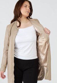 Beige läderjacka med en slät yta, öppen dragkedja och satinliknande foder. Modell som bär en vit topp och svarta byxor.