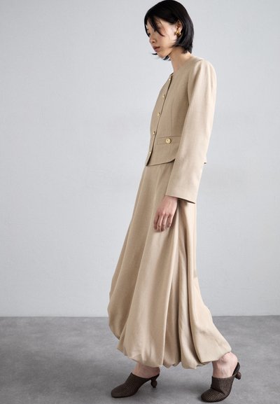 Ensemble beige en deux pièces comprenant un blazer ajusté avec des boutons dorés et une jupe fluide. Des mules en cuir tressé marron complètent l'ensemble.