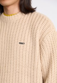 Suéter bege de malha com um padrão texturizado, gola canelada e um pequeno logo preto "OBEY" na parte da frente.