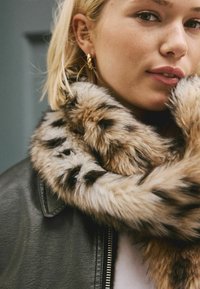 Écharpe en fausse fourrure dans des teintes de beige et de marron avec un motif tacheté, superposée à une veste en cuir noire, mettant en valeur une finition douce et texturée.