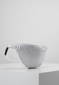 Sac banane en tissu gris avec une texture lisse, doté d'une sangle blanche et d'un logo rose, conçu dans une forme arrondie avec fermeture éclair.