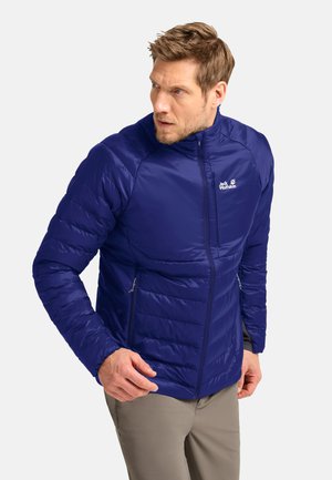Mann trägt eine blaue gesteppte Jacke mit vorderen und Brust-Reißverschlusstaschen, schaut zur Seite und steht vor einem schlichten Hintergrund.