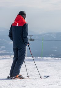 Skijacke mit rotem oberen Abschnitt und navyfarbenem unteren Teil, kombiniert mit navyfarbenen Hosen. Skier und Stöcke ruhen auf einem schneebedeckten Hang, im Hintergrund sind Berge sichtbar.