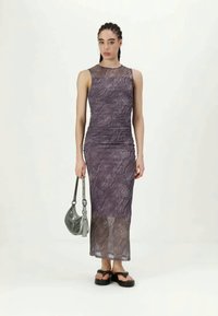 Μωβ φόρεμα χωρίς μανίκια με κυματιστό μοτίβο, εφαρμοστή σχεδίαση και διαφανή στριφώνα. Το μοντέλο κρατά μια γκρίζα τσάντα και φορά μαύρα σανδάλια.