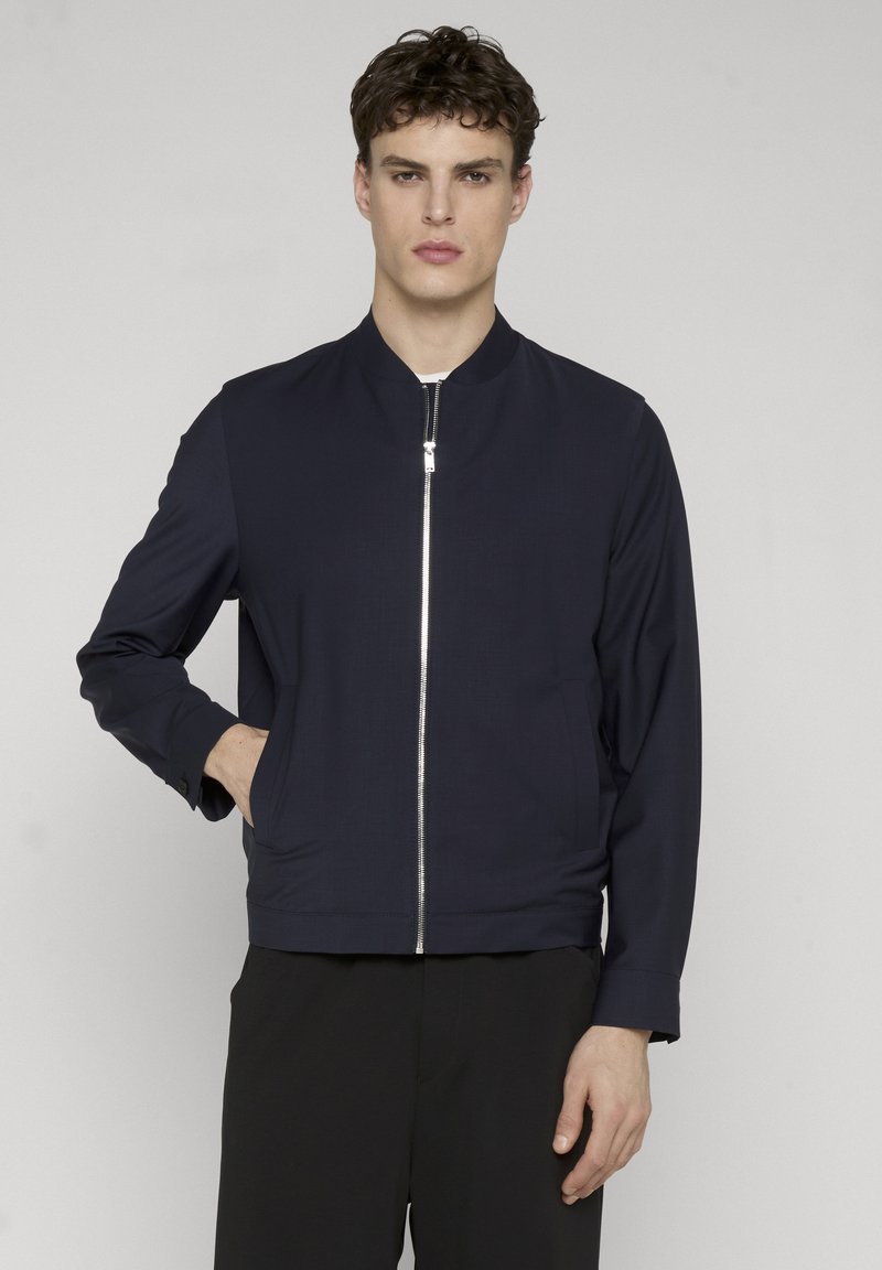 Giovane uomo con capelli ricci che indossa una giacca bomber blu navy con cerniera frontale, in piedi davanti a uno sfondo chiaro e semplice.