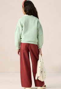 Un sweatshirt vert menthe avec des manches bouffantes, associé à un pantalon large rougeâtre. Un sac à dos crème avec un motif en forme de cœur est tenu dans une main.