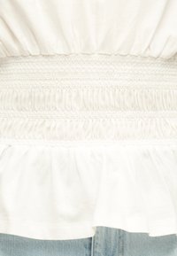 Tela blanca con bandas elásticas fruncidas horizontales y dobladillo con volantes fruncidos sobre mezclilla azul clara.