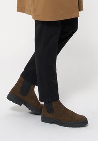 Brązowe zamszowe botki za kostkę z elastycznymi bocznymi panelami, w połączeniu z czarnymi spodniami. Botki mają teksturowaną powierzchnię i czarną gumową podeszwę.