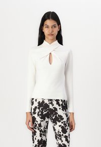White long-sleeve top with a twisted neckline and keyhole detail, worn with high-waisted black-and-white patterned pants.