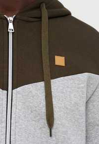 Φούτερ με κουκούλα δύο αποχρώσεων, με άνω μέρος σε ελιά πράσινο, κάτω μέρος σε ανοιχτό γκρι, κορδόνι σε ελιά πράσινο, ασημένιο φερμουάρ και μικρό τετράγωνο ταμπελάκι σε ταμπά στο στήθος.