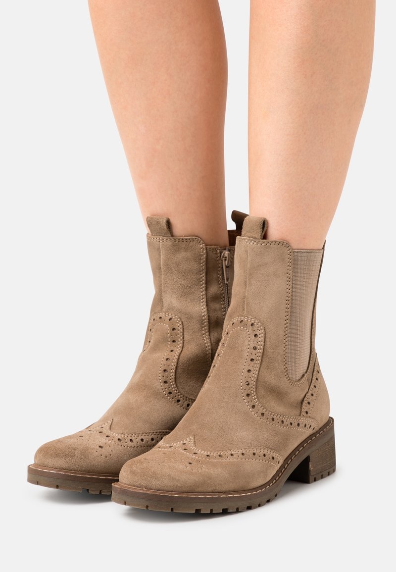 Bottes en daim couleur tan montant à la cheville avec panneaux latéraux élastiques, détails brogue décoratifs, talons blocs bas, portées sur jambes nues.