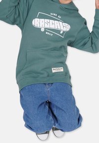 Grüne Kapuzenjacke mit "BAND OF RASCALS" Grafik, Fronttasche und umgestülpten Ärmeln; kombiniert mit lockeren blauen Denim-Jeans und schwarzen Sneakers.