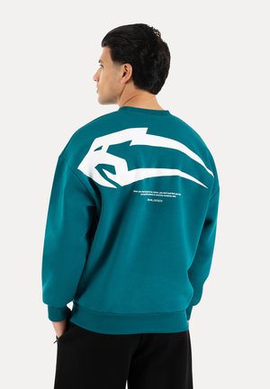 Man die van achteren is gezien, draagt een turquoise trui met een grote witte gestileerde grafiek en kleine tekst op de bovenrug, zwarte broek, handen in de zakken.