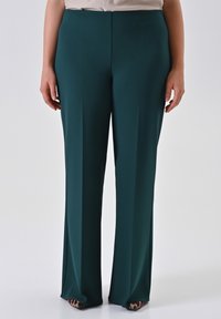 Persoon in hoge taille, wijde donkere groene broek en puntige schoenen met luipaardprint, staand tegen een effen lichte achtergrond.