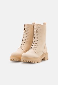 Bottes beige à lacets avec une semelle en caoutchouc épaisse, une texture lisse, un col rembourré et une languette; présentant un design robuste et un embout arrondi.