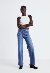 Lightweight white sleeveless top paired with high-waisted, wide-leg blue jeans. Black ankle boots complete the outfit. Simple, clean design.