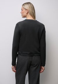 Svart, slim-fit sweatshirt med långa ärmar och rundad halsringning; texturerad tyg och elastisk fåll. Bärs med grå byxor.