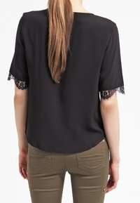 Svart blus med korta ärmar som har spetsdetaljer vid ärmsluten, slät tygkvalitet och rund halsringning. Kombination med khakifärgade byxor.