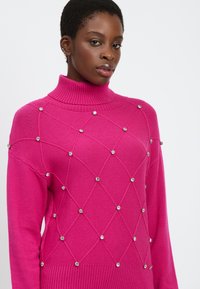 Fuchsia Strickpullover mit hohem Rollkragen, Rauten-Muster und Kristallverzierungen. Mit gerippten Bündchen und Saum.
