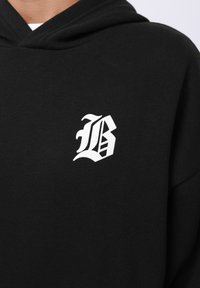 Hoodie negru dintr-un material moale, având un logo stilizat de culoare albă în formă de literă 'B' pe partea stângă a pieptului; glugă cu șnur și croială lejeră.