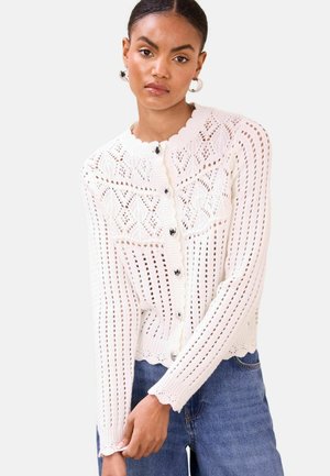 Mladá žena v bielom pletenom kardigáne so zapínaním na gombíky v tvare srdca a modrých džínsoch na jednoduchom pozadí.