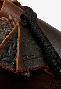 Scarpa in pelle marrone con finitura testurizzata, un laccio intessuto nero e il logo stampato "Clarks ORIGINAL" con un bordo decorativo.