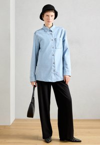 Ανοιχτό μπλε τζιν πουκάμισο σε oversized στιλ με κλείσιμο από κουμπιά και μπροστινή τσέπη, συνδυασμένο με μαύρες παντελόνες σε φαρδύ στιλ και μαύρο πλεκτό καπέλο τύπου bucket.