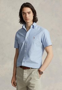 Junger Mann mit schulterlangem dunklem Haar, trägt ein hellblaues kurzärmeliges Hemd und beige Hose mit braunem Gürtel und Uhr.