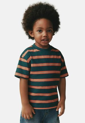 Niño pequeño con cabello rizado, vistiendo una camiseta de rayas verdes y naranjas y jeans azules, de pie sobre un fondo liso.