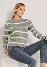Gestrickter Pullover in Hellgrau mit weißen Streifen, Rundhalsausschnitt und gerippter Textur. Kombiniert mit dunkelblauen Jeans und auffälligen gemusterten Accessoires.