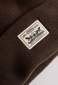 Berretto a coste marrone con etichetta rettangolare bianca contenente grafiche e testo nero, che indica il marchio di qualità Levi Strauss & Co.