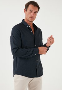 Camicia button-up blu scuro con maniche lunghe, colletto aperto e bottoni bianchi. Il tessuto sembra testurizzato, indossata con pantaloni chiari.