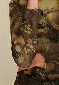 Gennemsigtig top med blomsterprint i dæmpede nuancer. Har flæseærmer og detaljer, der fremhæver indviklede mønstre af blomster og blade.