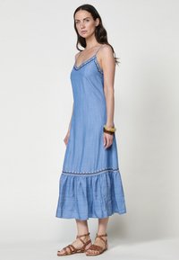 Robe d'été en lin bleu avec des bretelles spaghetti ajustables, un décolleté festonné et un ourlet à volants. Présente des coutures décoratives sur le buste.