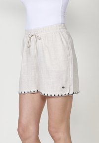 Shorts en lin beige clair avec une taille à cordon. Ils sont dotés d'un bord brodé noir le long de l'ourlet et d'un détail subtil de logo sur le côté.