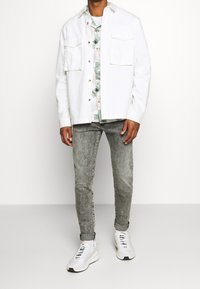 Camisa de mezclilla blanca con dos bolsillos frontales, llevada sobre una camisa de estampado floral en tonos pastel, combinada con jeans grises claros y zapatillas blancas.