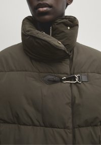 Olijfgroene pufferjack met een hoge kraag, voorzien van een metalen rits en zwarte leren accenten voor het vastmaken. Zachte, gewatteerde textuur.