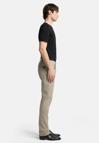 En person står på sidan iklädd en åtsittande svart t-shirt och beiga slim-fit byxor, kompletterat med svarta ankelstövlar.