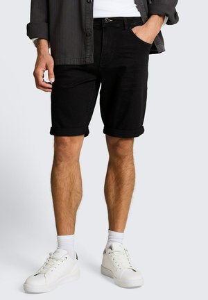 Man draagt opgerolde zwarte denimshorts, witte sneakers, witte sokken, wit overhemd en donkergrijze jas met één hand in de zak.