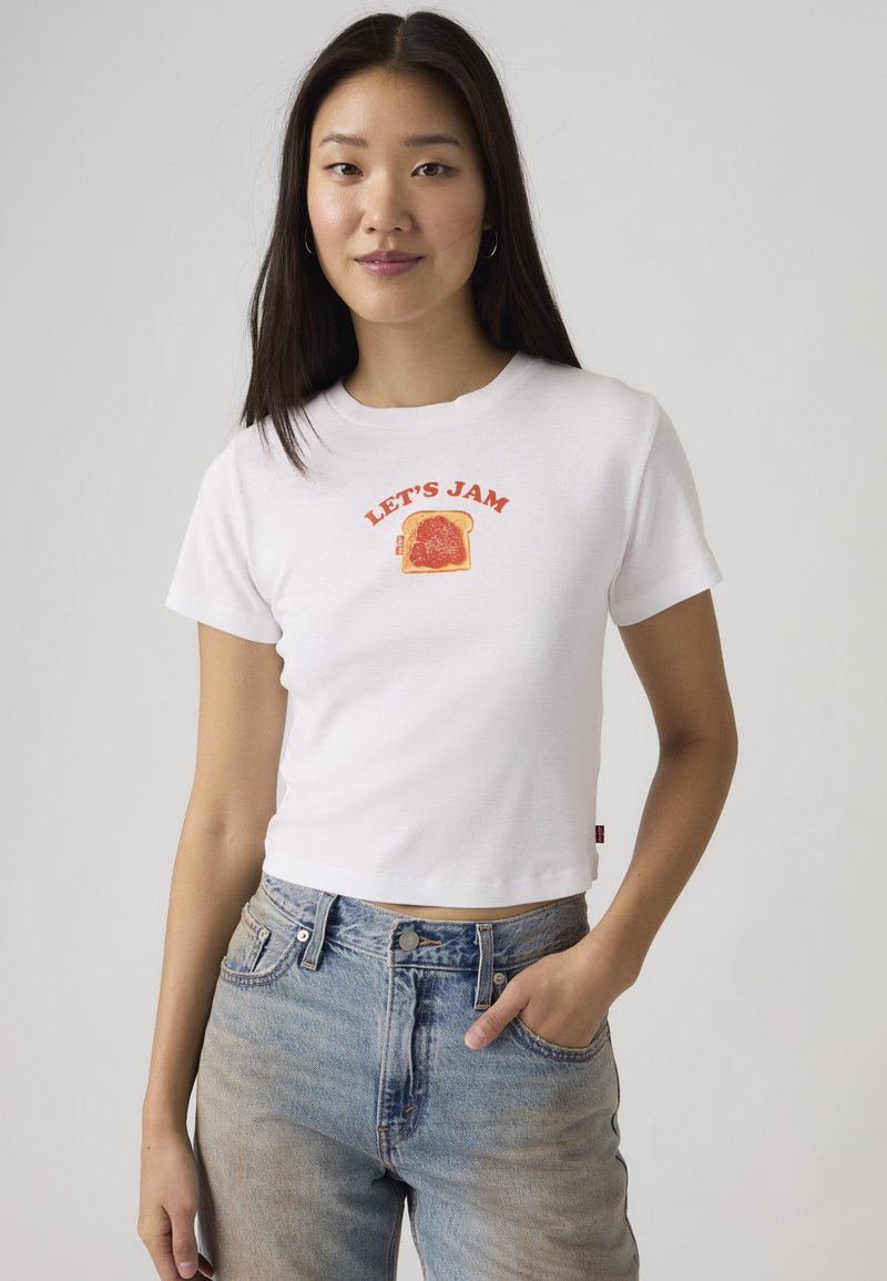 Νεαρή γυναίκα με ίσια σκούρα μαλλιά φοράει ένα λευκό crop t-shirt με την επιγραφή «LET'S JAM» πάνω από μια φέτα ψωμί με κόκκινη μαρμελάδα, συνδυασμένο με ανοιχτόχρωμα μπλε τζιν.