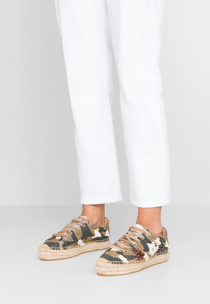 Pieds portant un pantalon blanc court et des baskets à lacets avec un imprimé camouflage, des paillettes dorées et une semelle espadrille tissée.