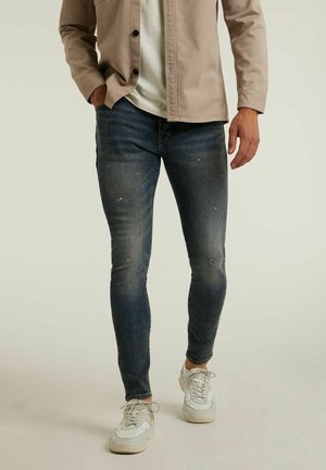 Jean skinny en denim bleu foncé délavé, avec des détails usés et une coupe ajustée. Porté avec une chemise de couleur claire et des baskets blanches.