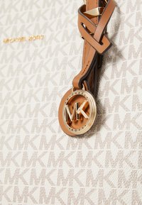 Tote bag blanche avec un motif répétitif "MK", agrémentée d'une étiquette de logo circulaire dorée. Caractéristiques d'accents en cuir marron et d'une surface texturée.