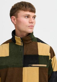 Fleece-pullover i en lapptäcktsdesign av grönt, brunt och gult. Har en ficka med dragkedja på bröstet och hög krage. Mjuk textur.