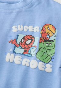 Vaaleansininen puuvillatakki, jossa on graafisia printtejä Hämähäkkimiehestä, Rautamiehestä ja Hulkista, ja jossa on kohotettu valkoinen teksti "SUPER HEROES."