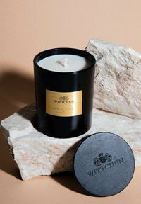 Vaso di candela nera con etichetta dorata, cera bianca e stoppino in cotone. Accompagnato da un coperchio in legno nero inciso con "WITTCHEN." Appoggiato su una pietra texturizzata.