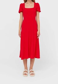 Punainen midi-mekko lyhyillä puffihihnoilla, neliökaula-aukolla, istuvalla bodysuunnittelulla ja valuilla hameilla. Yhdistetty valkoisten sandaalien kanssa. Sileä kankaan tekstuuri.