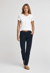 Hvid v-hals t-shirt med lille sort logo, mørkeblå bukser, brun bælt og beige mokkasiner. Model står op ad en ensfarvet baggrund.