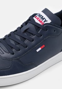 Sneaker in pelle blu con tomaia testurizzata, punta rotonda e suola in gomma bianca. La linguetta con il marchio mostra "TOMMY JEANS" e accenti con la bandiera colorata.